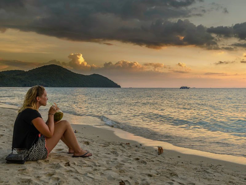Vrouw zit op een zandstrand met kokosnoot in de hand en kijkt uit over zee bij zonsondergang, met een eiland en boot in de verte.