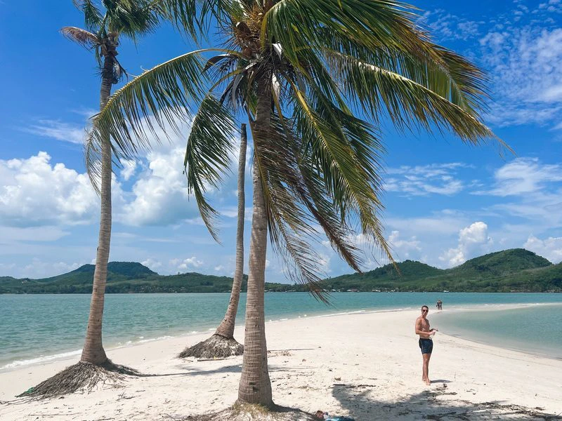Man staat op een smalle zandstrook tussen turquoise zee en palmbomen, met heuvelachtig eiland op de achtergrond en een blauwe lucht vol wolken.