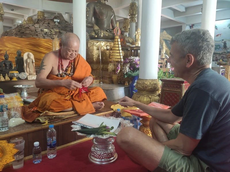 In een kleurrijke tempel zit een boeddhistische monnik in oranje gewaad tegenover een reiziger. De monnik bindt een rood armbandje om zijn pols tijdens een zegenceremonie. Op de achtergrond staan beelden en gouden versieringen.