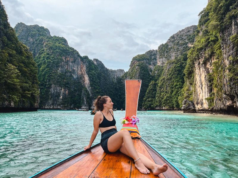 Reizigster zit ontspannen op een traditionele longtailboot in het heldere turquoise water van een kalkstenen baai in Zuid-Thailand, omringd door groene rotsformaties en tropisch landschap.