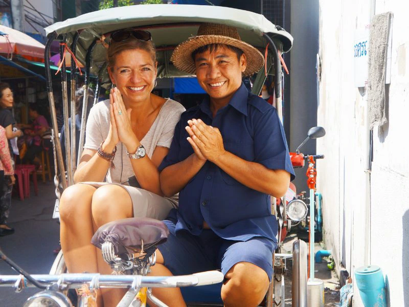 Reizigster lacht samen met een vriendelijke Thaise tuktuk-chauffeur in een smalle straat van Bangkok, beide maken een traditionele Thaise begroeting met gevouwen handen terwijl de lokale sfeer zichtbaar is op de achtergrond.
