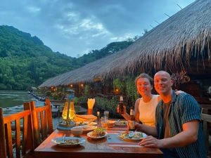 Vrouw en man die lachend in de camera kijken. Ze zitten bij de rivier bij Kanchanaburi te dineren. Je ziet de houten drijvende huisjes op de achtergrond.