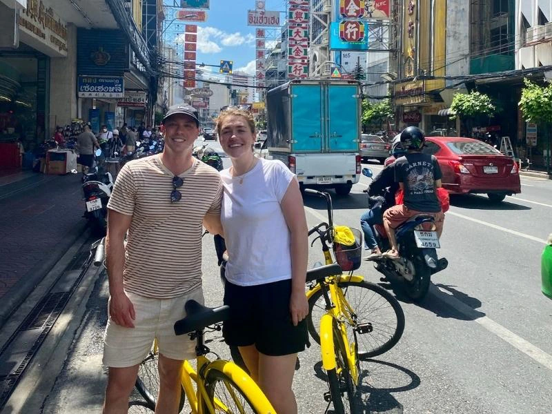 Man en vrouw staand bij hun gele fietsen in Chinatown in Bangkok. Ze lachen naar de camera. Ze staan in een drukke straat met veel verkeer.