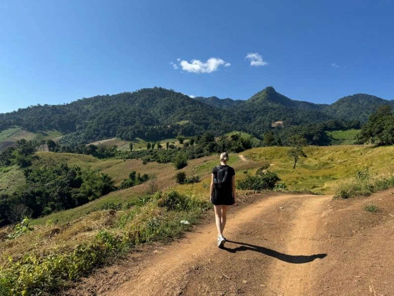 Vrouw met een rugtas op wandelend door de heuvels van Noord-Thailand. De omgeving is groen en de lucht is blauw.