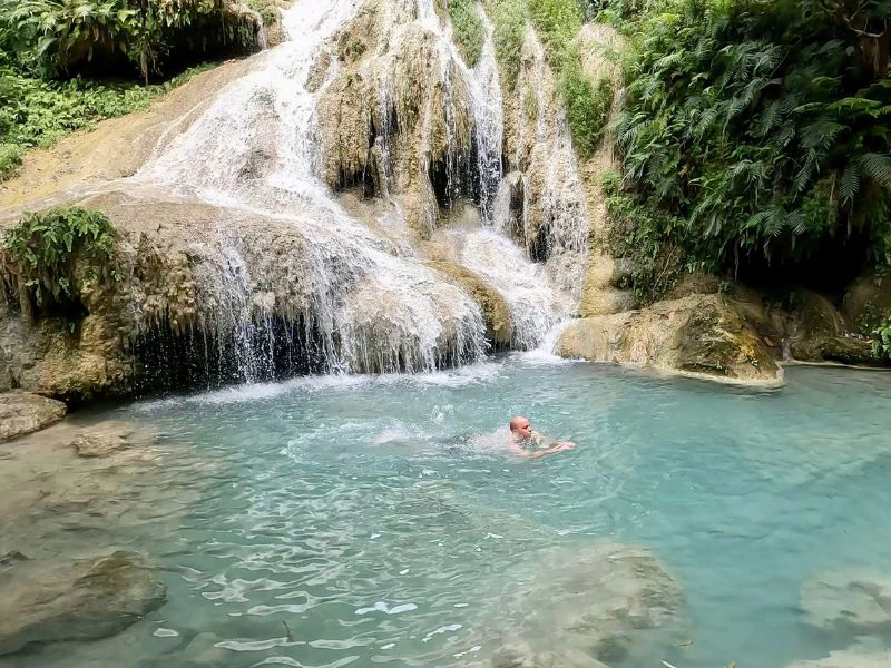Man zwemt in een helderblauw natuurpoeltje aan de voet van een kleine waterval, omringd door rotsen en groen.