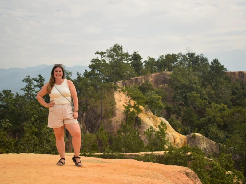 Reizigster met hand boven haar heup staat lachend te poseren op de zanderige en kronkelende Pai Canyon. Met diepe afgronden en veel groen omringd. De lucht is grijzig.
