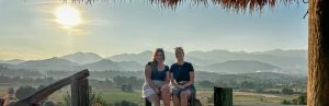 Twee vrouwen zitten lachend op een houten bankje met uitzicht op groene rijstvelden en bergachtig landschap bij ondergaande zon.