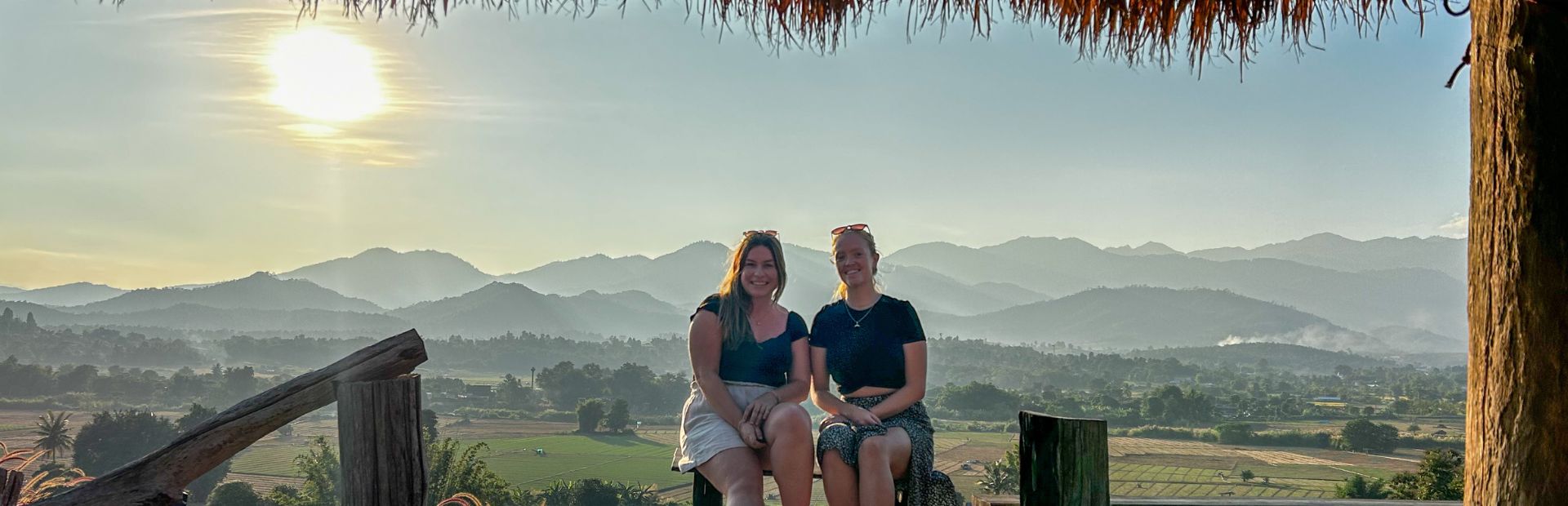 Twee vrouwen zitten lachend op een houten bankje met uitzicht op groene rijstvelden en bergachtig landschap bij ondergaande zon.