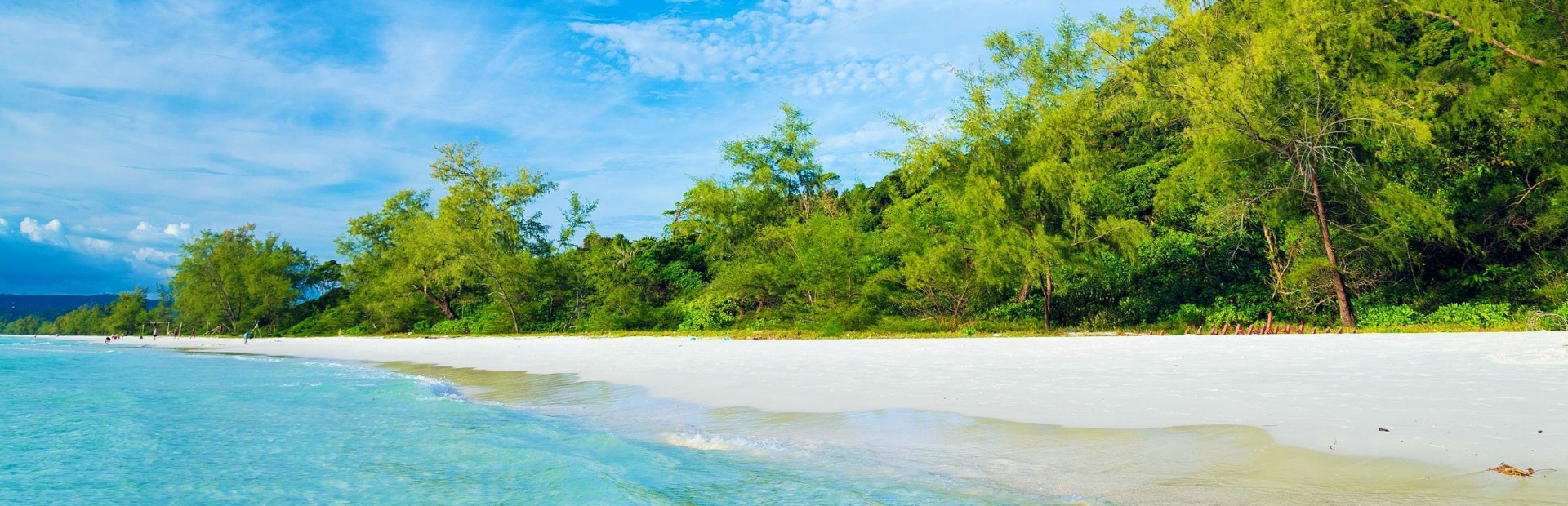 het witte lange strand van Koh Rong, omzoomd door groene palmbomen met op de voorgrond een lichtblauwe kabbelende zee