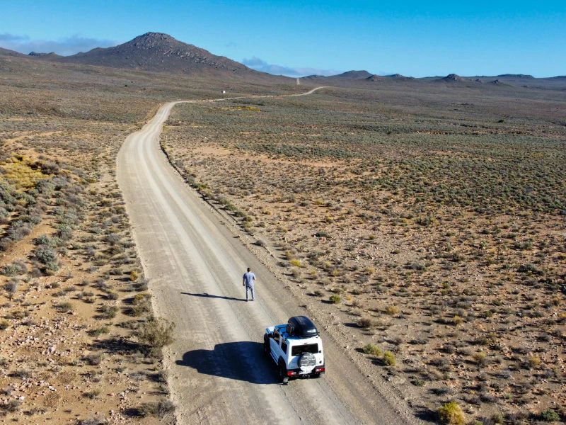 Man staat op een weg in Zuid-Afrika voor zijn auto. Het landschap is vrijwel leeg en in de verte zie je bergen