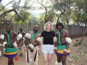 Een groep jonge mannen in traditionele Swazi-kleding, gekleed in kleurrijke rokken en versierde armbanden, poseert buiten naast een vrouw in een zwart T-shirt en gestreepte korte broek. Ze staan samen voor een houten hek, omringd door bomen en groen.