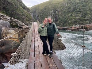 Twee jonge meiden staan op een houten hangbrug boven het wilde water en tussen rotsen met veel groen erop.
