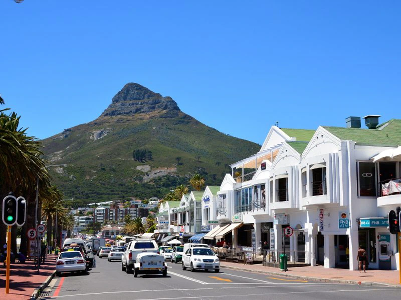 Een levendige straat in Kaapstad met witte gebouwen, restaurants en winkels aan de rechterkant. Er rijden auto's en taxi's op de weg, en er zijn verkeerslichten en palmbomen langs de straat. Op de achtergrond is een groene berg met een karakteristieke piek te zien onder een heldere blauwe lucht.
