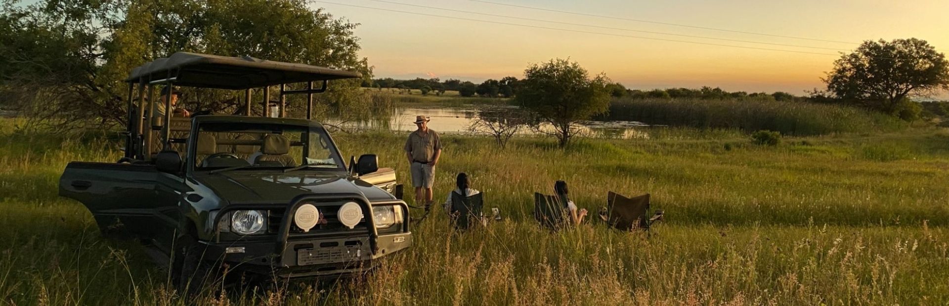 Drie mensen op een stoeltje kijken uit over een meer in Limpopo. Een man staat naast hen en daarnaast staat de safari auto.