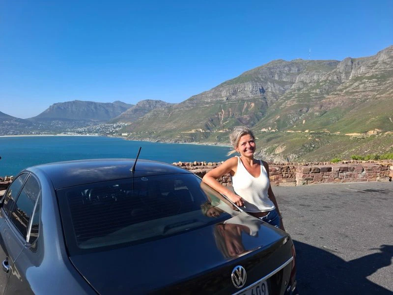Vrouw staat tegen een zwarte auto te leunen bij een uitzichtpunt langs de weg met uitzicht op de blauwe zee, groene rotsachtige hoge bergen en de stad in de verte. De lucht kleurt helderblauw.