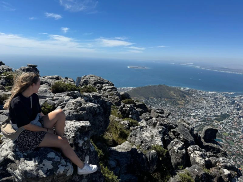 Een vrouw zit op rotsen bovenop een berg en kijkt uit over de stad en de oceaan beneden. Ze draagt een donker T-shirt, een kort broekje met zwart-wit patroon en witte sneakers. Haar haar waait in de wind. Voor haar liggen grote grijze rotsformaties en daarachter is een weids uitzicht over Kaapstad, de kustlijn en een helderblauwe zee onder een blauwe lucht met enkele wolken.
