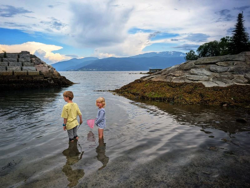 Twee kinderen staan met laarzen aan in het water aan een strandje.
