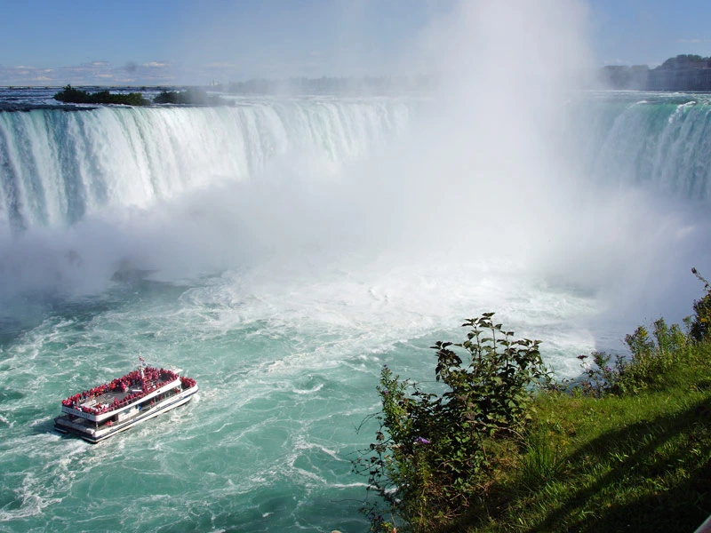 Rondvaartboot vol toeristen vaart dicht langs de imposante Niagara Falls, waar grote watermassa’s met kracht naar beneden storten en nevel omhoog spuit.