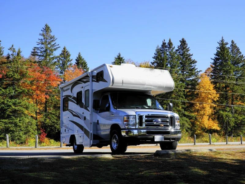 Witte camper geparkeerd langs de weg met kleurrijke herfstbomen op de achtergrond.