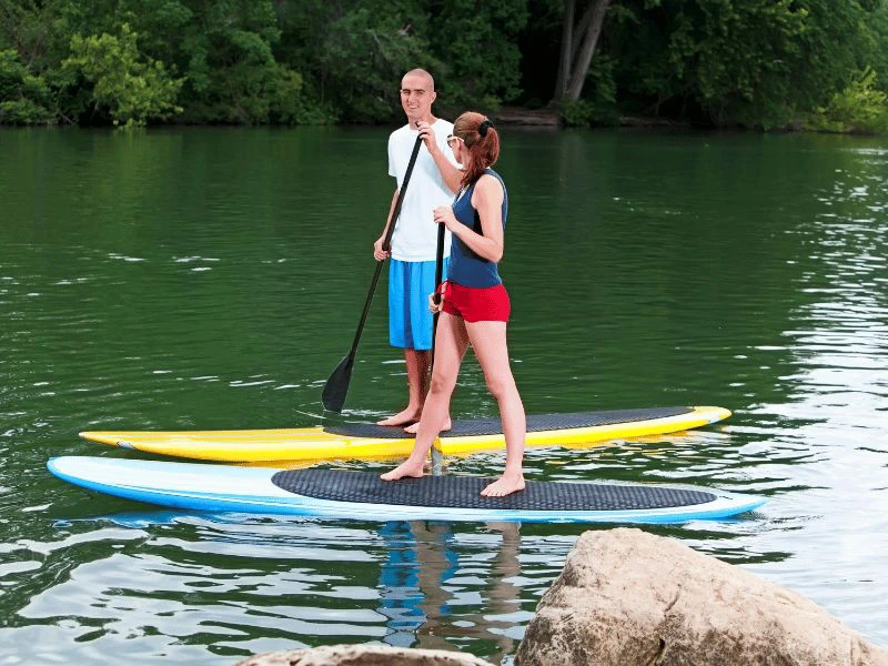 Een man en een vrouw staan op paddleboards en peddelen rustig over een groen meer met bomen langs de oever.
