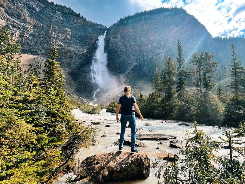 Reiziger staat op een rots in een stromende rivier met bergen en een waterval op de achtergrond.