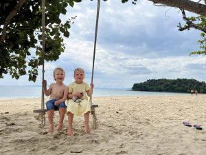twee kleine kinderen zitten op een schommel die aan een boom hangt die boven in de foto het beeld in komt. Het strand is leeg. In de verte is de zee en daarachter een klein bebost eiland. De lucht is bewolkt.