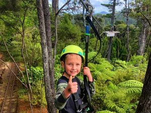 Kind met groene helm en klimgordel geeft een duim omhoog terwijl hij vastzit aan een zipline, omringd door groene bomen en varens; op de achtergrond een platform tussen de bomen.