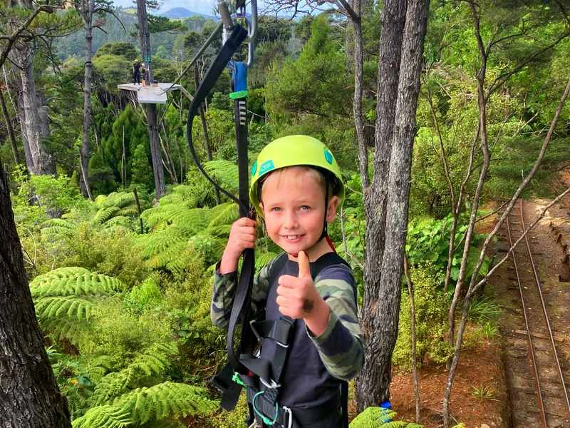 Kind met groene helm en klimgordel geeft een duim omhoog terwijl hij vastzit aan een zipline, omringd door groene bomen en varens; op de achtergrond een platform tussen de bomen.