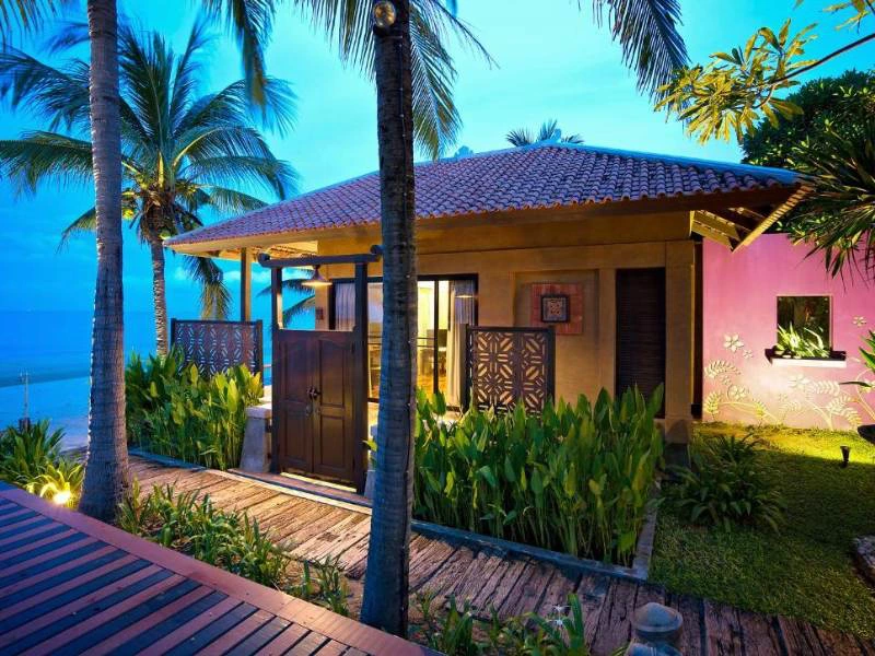 Sfeervol verlichte strandbungalow omgeven door palmbomen en tropische planten tijdens de schemering.