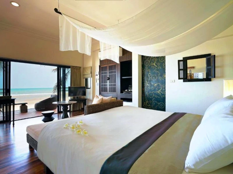 Luxe hotelkamer met hemelbed en uitzicht op het strand en de zee via open schuifdeuren.