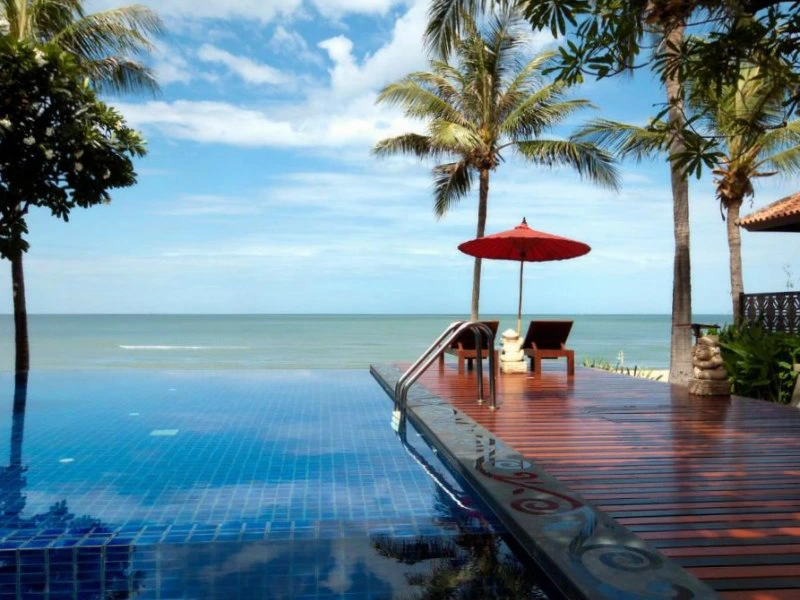 Infinity pool met houten terras, rode parasol en uitzicht op zee en palmbomen.