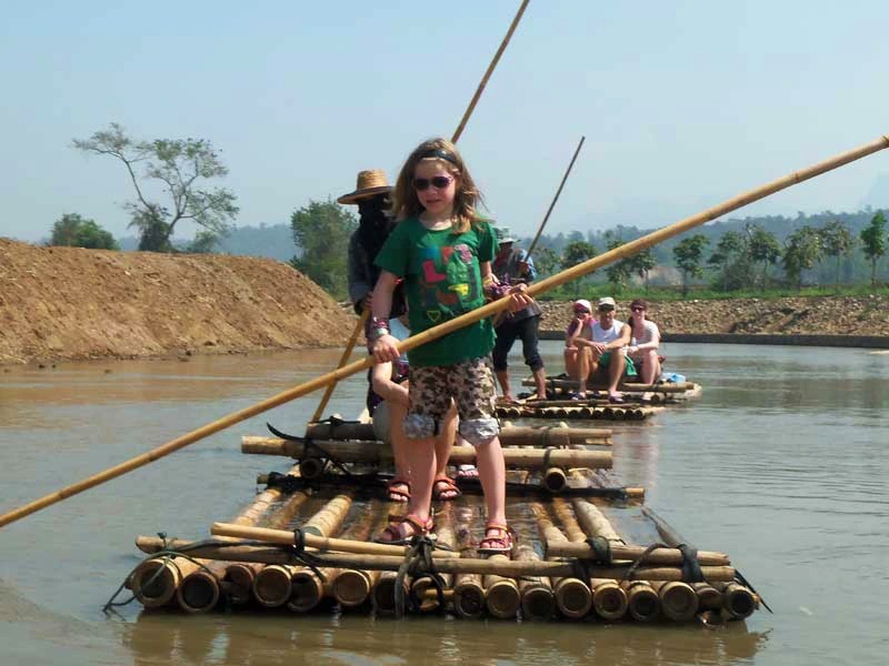 Een meisje met een zonnebril staat met een bamboestok op een bamboevlot op een rustige rivier; op de achtergrond zitten andere mensen op een tweede vlot.