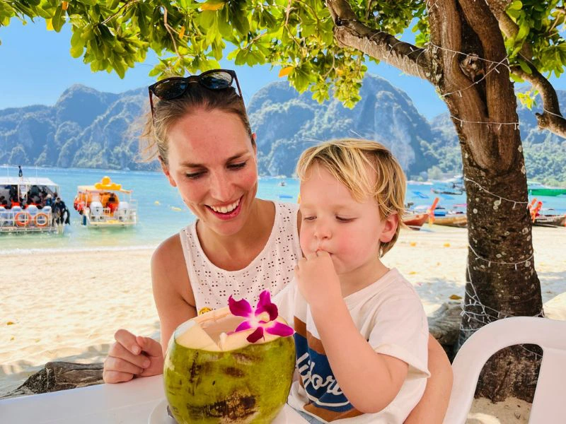 Vrouw met kind drinken uit een kokosnoot met strand en bergen op de achtergrond