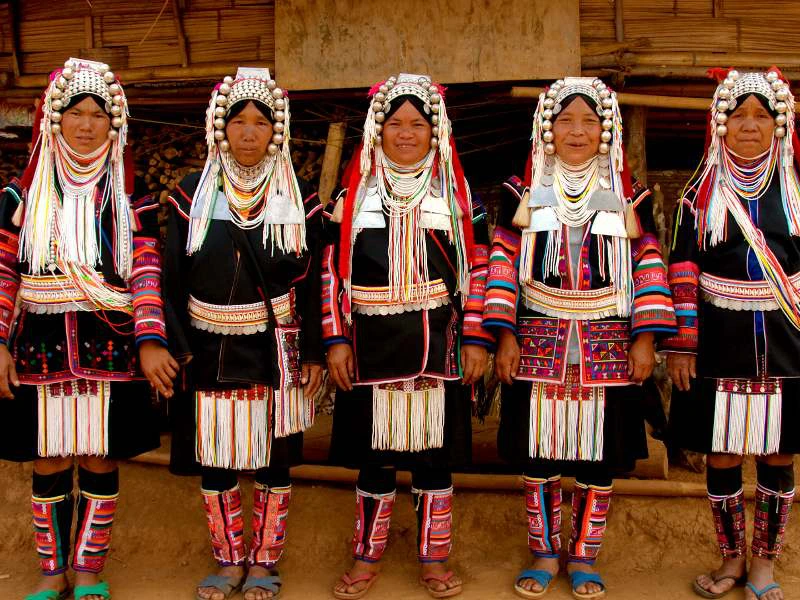 Vijf vrouwen in kleurrijke traditionele klederdracht met hoofdtooien, kralen en borduursels, poserend voor een houten gebouw.