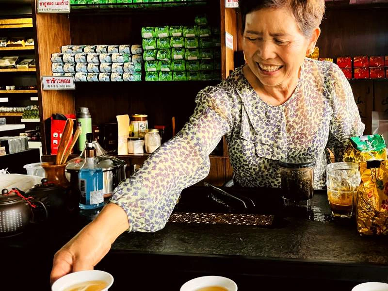 Glimlachende vrouw serveert thee in een traditionele theewinkel, met kleurrijke verpakkingen en theepotten op de achtergrond.
