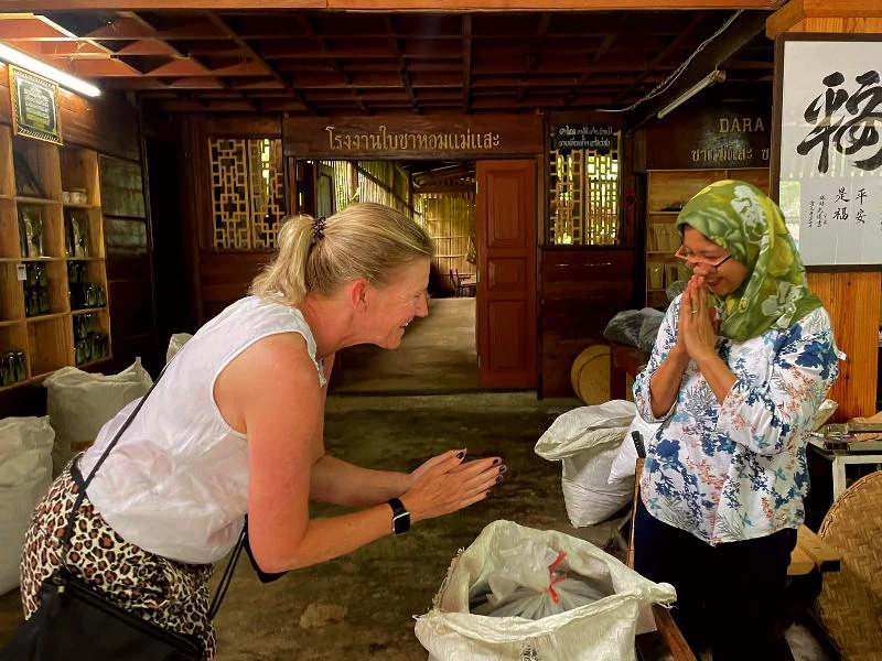 Twee vrouwen groeten elkaar met gevouwen handen in een traditionele houten ruimte, omringd door zakken en rekken met theeverpakkingen.