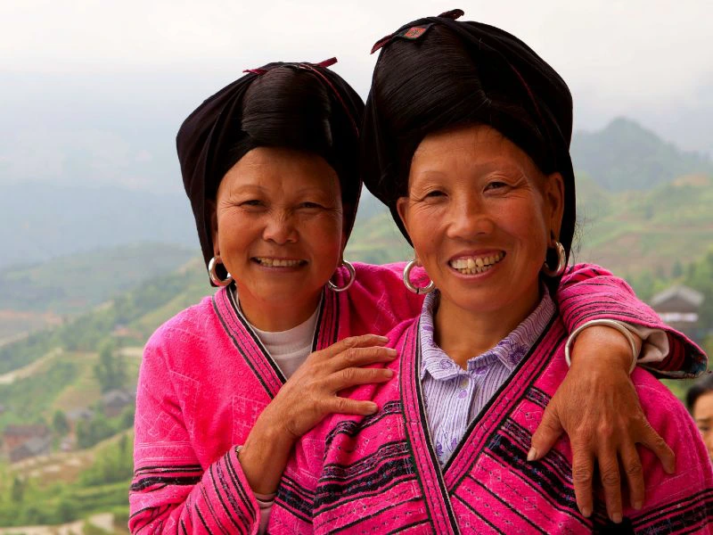Twee vrouwen in felroze traditionele kleding en zwarte hoofddeksels, met grote zilverkleurige oorringen, poseren glimlachend voor een heuvelachtig landschap.