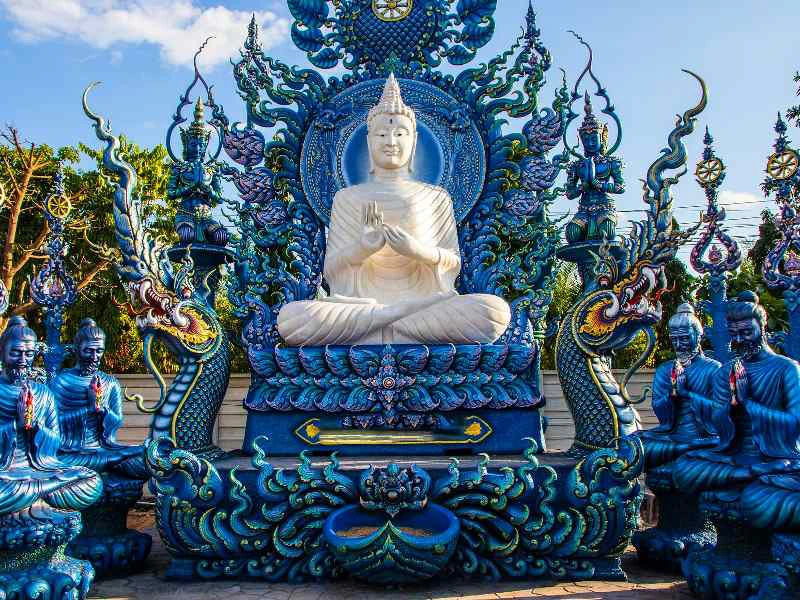 Witte Boeddhabeeld op rijkelijk versierde blauwe troon met sierlijke naga-figuren en mediterende beelden eromheen.