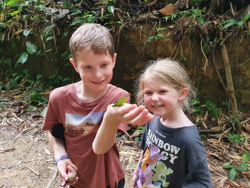 Twee kinderen bekijken een groene vlinder op de hand van een jongen in een bosrijke omgeving.