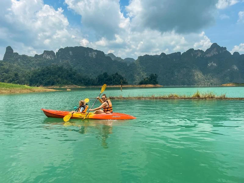 Twee mensen in een oranje kajak op helder turquoise water met jungle en karstrotsen op de achtergrond.