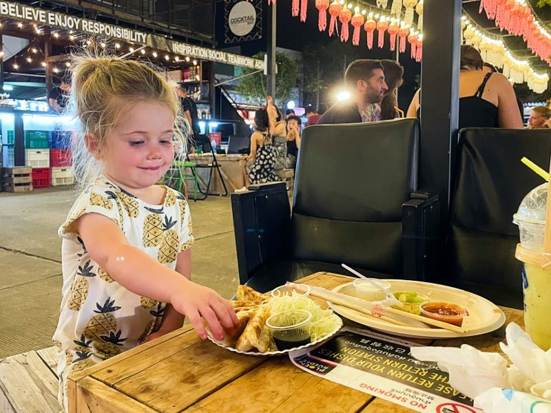 Klein meisje eet streetfood op een levendige avondmarkt.