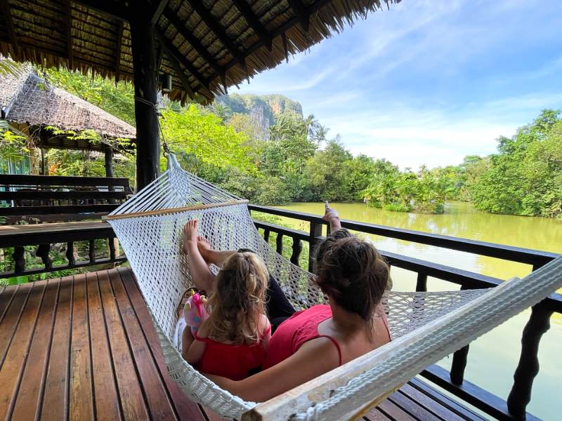 Moeder en dochter liggen samen in een hangmat op een houten veranda met uitzicht op een groene rivier en tropische natuur.
