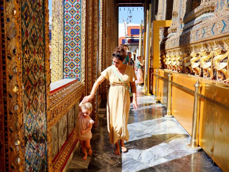 Vrouw met jong kind loopt langs rijkversierde gouden muur in een tempel in Thailand, badend in het zonlicht