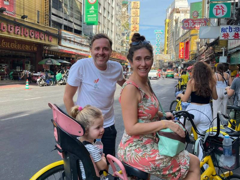 Gezin op fietsen in drukke straat met Aziatische winkels en kleurrijke uithangborden.