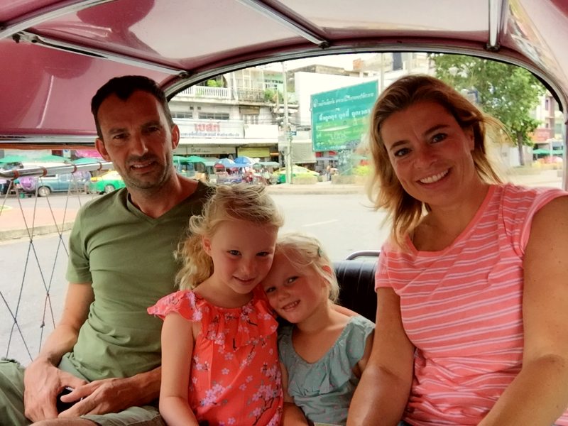 Gezin van vier zit glimlachend in een open tuk-tuk in een drukke stad.