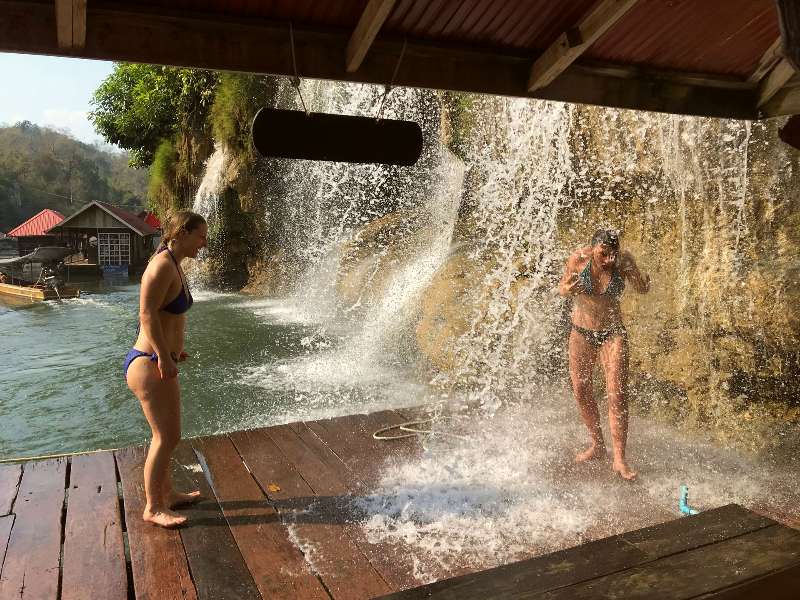 Twee vrouwen in bikini onder een waterval op een houten vlonder aan het water.