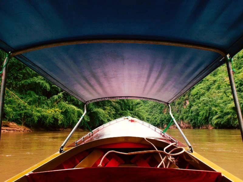 Uitzicht vanuit een longtailboot met blauw dak op een bruine rivier tussen groene beboste oevers.