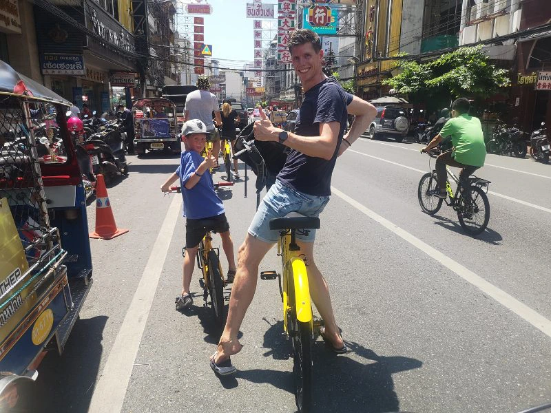 Links zoon en rechts vader op gele fietsen door Chinatown in Bangkok. Voor hen nog meer fietsers van dezelfde tour. Beide draaien even om en steken hun duim op. Je ziet de borden van Chinatown langs de gebouwen en om hen heen een tuktuk en andere weggebruikers.