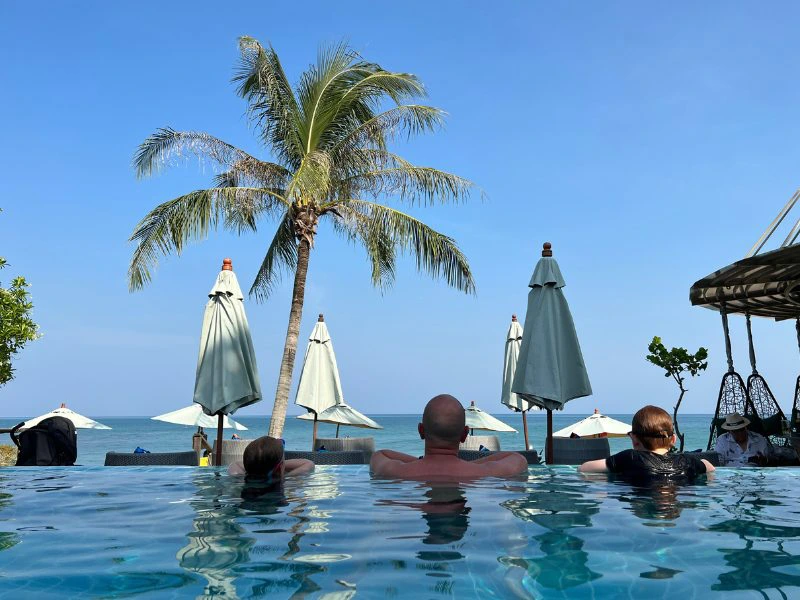 Vader met twee kinderen hangen over de rand van het zwembad met ruggen naar de camera, kijkend naar de dichte parasollen en palmbomen en de helderblauwe lucht.