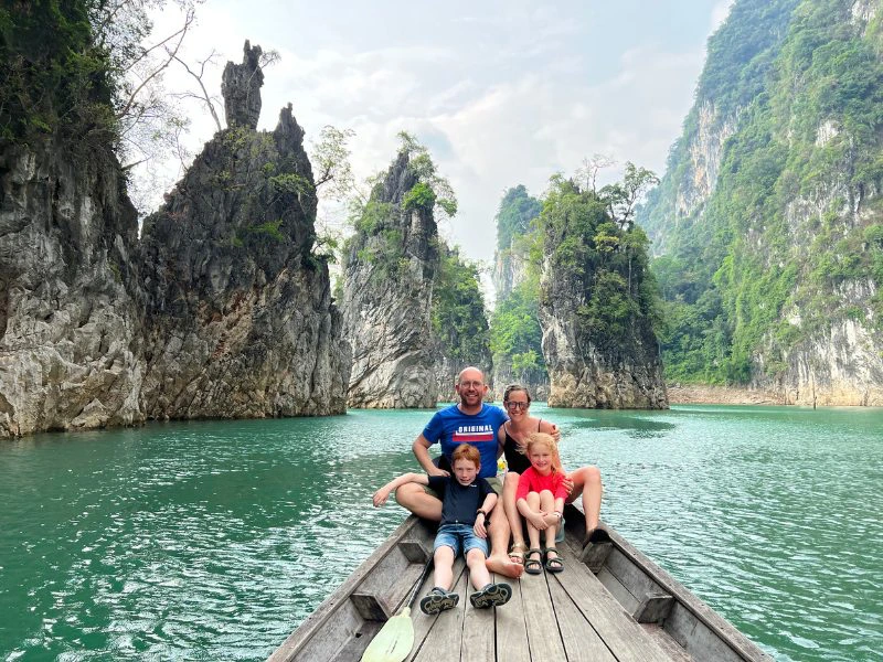 Een gezin van vier zit voorop in een smal houten bootje op een helderblauw meer, omringd door hoge, steile kalkstenen rotsen en groene jungle in Khao Sok National Park, Thailand. De ouders zitten achterin met hun armen om de kinderen heen, die vooraan op de rand van de boot zitten. Iedereen kijkt lachend in de camera. Het water is kalm en de natuur om hen heen oogverblindend groen. De sfeer is ontspannen en avontuurlijk, typisch voor een gezinsreis door Thailand.